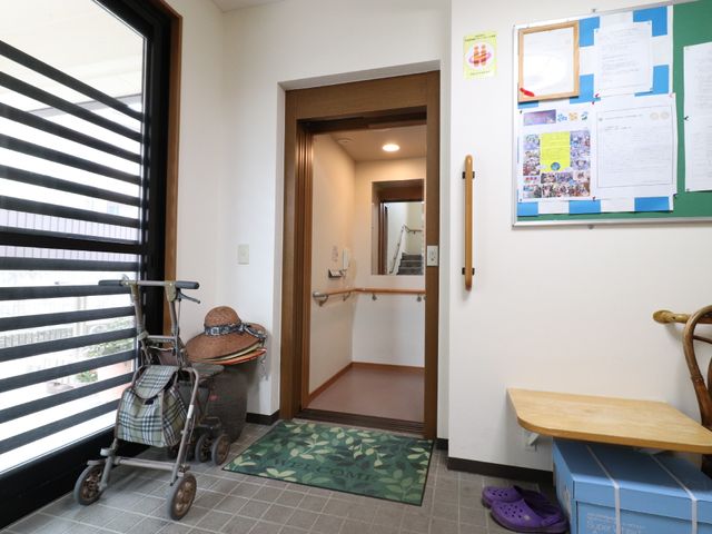 出入りしやすい開口部 エレベーター 手すりが配された入口には、歩行器が置かれており、入居者の安全と利便性を考慮した設計です。壁には掲示物があり、コミュニケーションの場も提供しています。