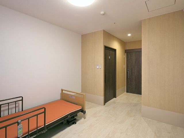 居室のベッドとドア 居室 きれいに整えられた居室で、明るく心地のよい空間を提供しています。安全を考慮したシンプルなベッドも配備されております。