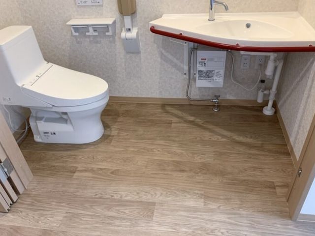 明るいトイレ トイレ   手すり付きの個室トイレで、洗面台も近くに備わっています。床は木目調で、清潔感のある印象です。