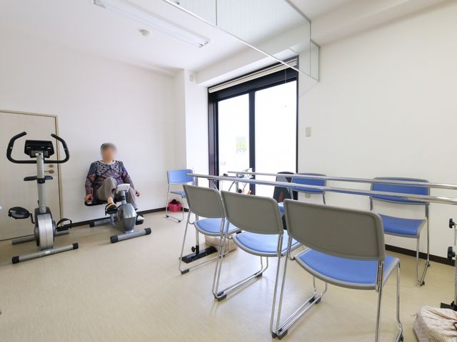 機能訓練室と利用者 共有スペース 清潔感のある機能訓練室で、一人の利用者がリハビリトレーニングを行っています。周りには安定した椅子も完備されており、支えが必要な方も安心です。