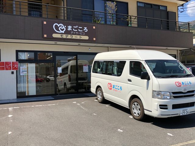 白い車が停まる建物前 外観 施設の前には、訪問車両が停まっており、アクセスの良さが魅力です。暖かい日差しを浴びるエントランスが安心感を与えます。