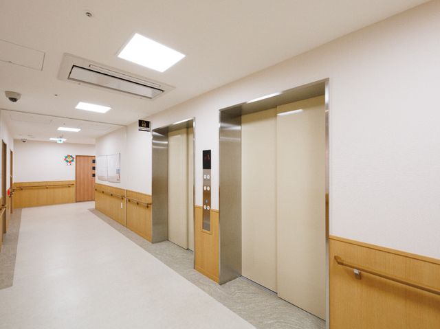 清潔な廊下のエレベーター エレベーター 清潔感のある廊下にはスムーズに動くエレベーターが設置され、職員や利用者様の移動をサポートしています。木目調の壁が温かみのある雰囲気を醸し出しており、安心してお仕事を始められる環境です。