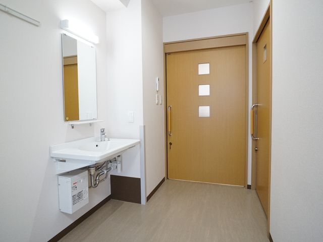 洗面台と入口の部屋 居室 清潔感あふれる居室の入口にはハンドル付きの扉が設けられ、安全かつ便利な生活環境が整っています。洗面台は室内に備え付けられており、身の回りの小さなお世話も容易です。