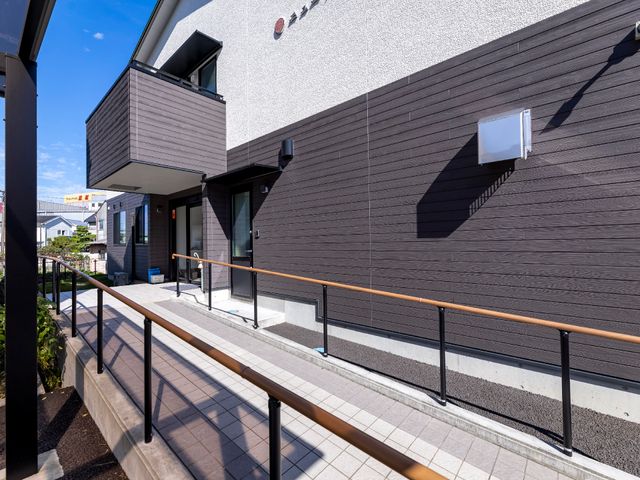 明るく清潔な施設のエントランス 玄関 手すり沿いのバリアフリー設計のエントランスは、訪れる方に安心を提供します。清潔感のある外観が印象的です。