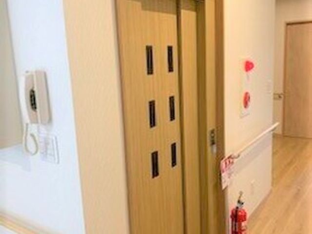 清潔感ある廊下と扉 エレベーター 手すり付きの廊下に臨む、透明感のあるガラス窓のエレベーターが設置されており、利用者の移動が容易になっています。