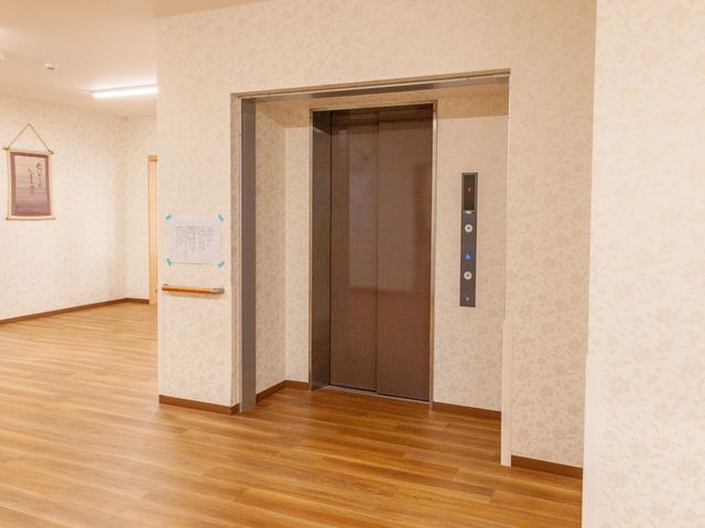 きれいな廊下のエレベーター エレベーター 床の木目と壁の柄がやさしい雰囲気の廊下です。掲示物が見やすい位置に掲げられ、利用者の方々の安全と情報提供に配慮されています。