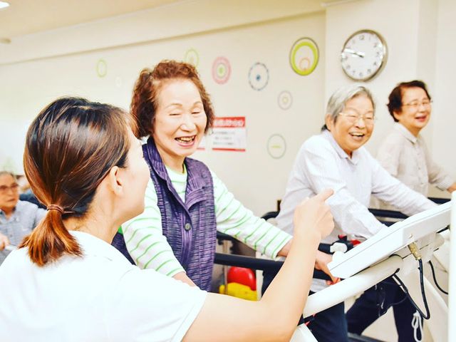 笑顔で機能訓練中の高齢者 活気に満ちた機能訓練室で、利用者が明るい笑顔で体を動かしています。職員のサポートも手厚く、心地よいコミュニケーションが育まれています。