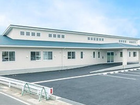 平屋建ての建物の外壁は白でグリーンの屋根が付いている。広い駐車場があり、日当たりのいい場所に建っている。