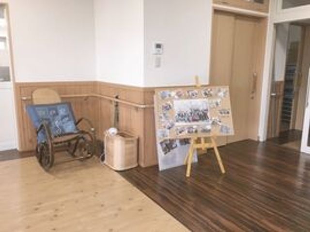 廊下に展示と車椅子 玄関 広々とした廊下が設計されていて、壁際にはアクティビティの写真が飾られ、訪れた方々に暖かい印象を与えます。ゆったりとした空間で、日々の生活が快適に過ごせそうです。