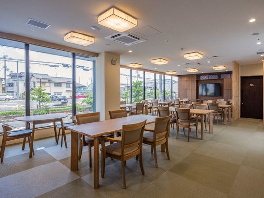施設の写真 大きな窓があり明るい雰囲気。中央にダイニングテーブルが複数置かれ、肘置き付きの椅子がセットされている。