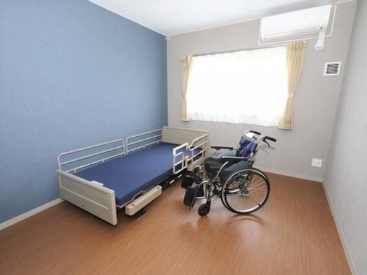 車いすや介護ベッドが置かれたフローリングの居室には、カーテン付きの窓もあり、壁は一部ブルー。