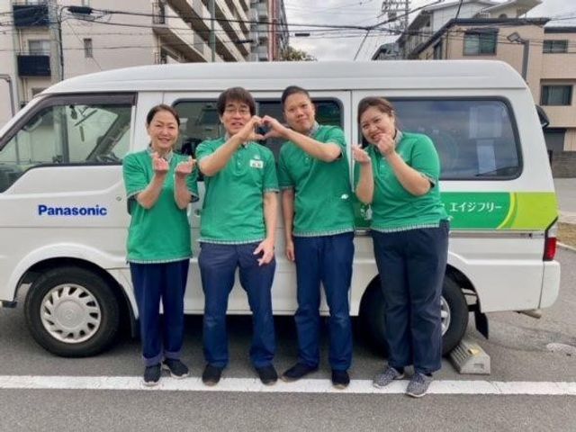 スタッフと送迎車 スタッフイメージ   緑色のユニフォームを着たスタッフの皆さんが、送迎車の前で笑顔を見せています。チームの雰囲気が伝わり、就業後の連携をイメージしやすい場面です。