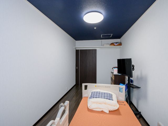 個室のベッドとテレビ 居室 シンプルで機能的な個人の居室です。必要な物をすぐ手に取れるよう、ベッドサイドには収納スペースがあります。