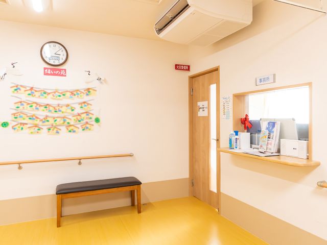 明るい受付空間 受付 暖かみのあるフロントは、待合いベンチも備え、訪れる人を優しく迎え入れます。壁に掲げられた時計下の季節の飾りが、明るく穏やかな雰囲気を演出しています。