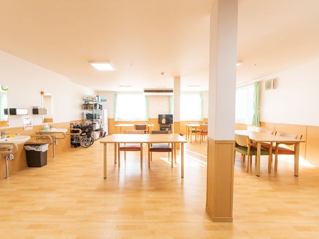 広々とした明るい居室 共有スペース 床が木目調で落ち着いた雰囲気の食事スペースがあり、車椅子が使われているのが見受けられます。ゆったりとくつろげる環境を大切にしています。