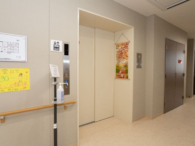 廊下のエレベーター前景 エレベーター 手すりが設置された清潔感のある廊下です。移動のサポートや手指消毒ステーションが整っており、安全と衛生を心がけた環境が提供されています。
