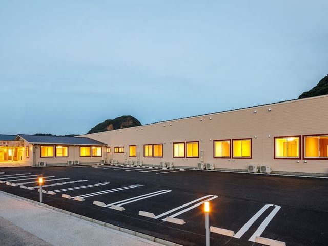 夕景に照らされた一階建て建物 外観 ゆったりとした駐車スペースを備え、夕暮れ時の温もりある灯りが窓から溢れる落ち着いた雰囲気の施設です。