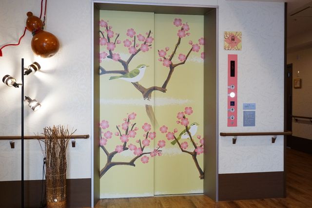 春を感じるエレベーター前 エレベーター 桜模様が施されたエレベーター前は、訪れる人々に穏やかな春の息吹を感じさせます。廊下に優しい灯りを与える照明が、空間に温もりを加えています。