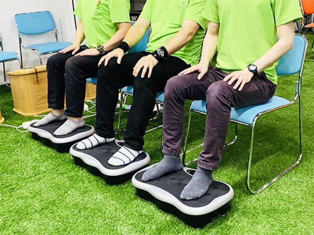 機能訓練中の様子 機能訓練室 利用者が椅子に座って、専用機器を使った足の運動に取り組んでいます。リラックスした雰囲気です。