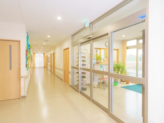 明るい廊下のある施設 玄関 ゆとりある通路は清潔で明るく、壁にはアートが飾られており、暖かな雰囲気が感じられます。ガラス窓越しに見える部屋も整っており、心地よい環境での就業が期待できそうです。