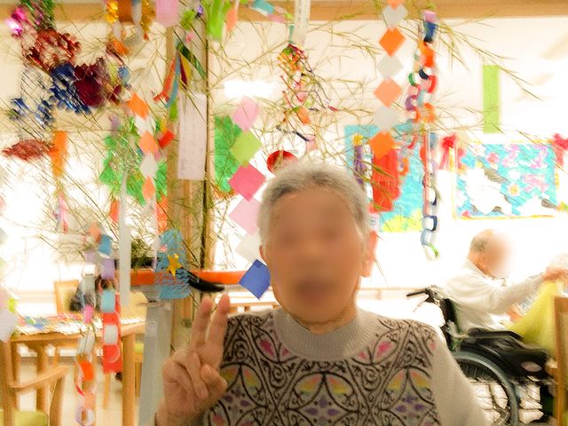笑顔のおばあ様と節分飾り ケア 節分を祝う装飾が施された室内で、おばあ様がピースサインをして明るく微笑んでいます。賑やかな雰囲気で日々の生活が楽しめそうです。