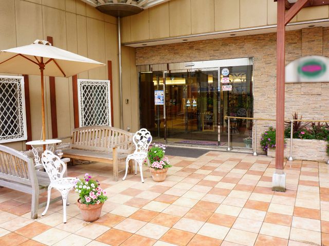 暖かみのあるエントランス 正面玄関 エントランスには木製のベンチと白いガーデンチェアが配置され、訪れる方を優しい雰囲気で迎えます。花の鉢植えが彩りを添え、安らぎの空間を演出しています。