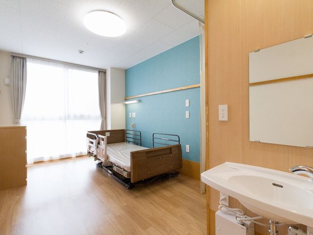 ベッドと洗面台の居室 居室 広々とした明るい居室で、床も清潔に管理されています。自然光が優しく差し込む環境です。