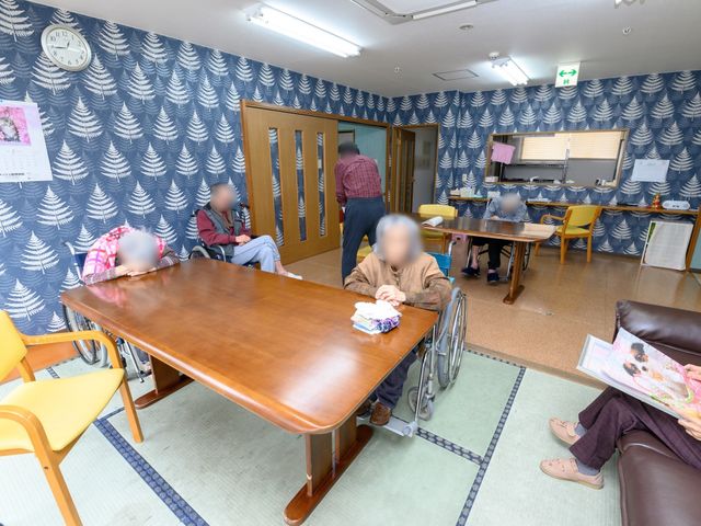 複数の高齢者がいる居室 リビング ゆったりとした木製のテーブルを囲み、高齢者がくつろいでいる暖かみのある部屋です。椅子に座る方や車椅子を利用されている方々の和やかな様子が伝わってきます。