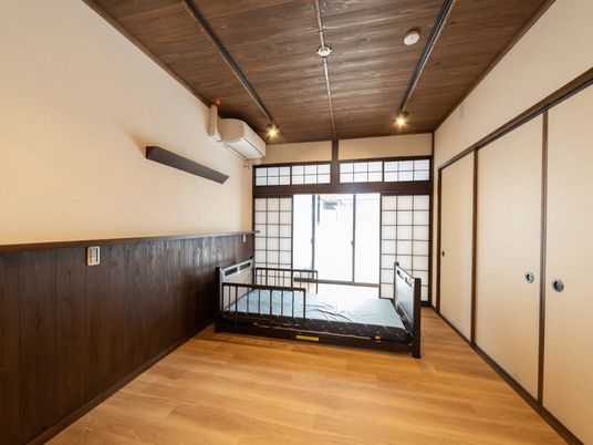 寝具のついていな介護ベッドがフローリングの部屋の中央に置かれ、壁にはエアコンが付き、大きな窓にはカーテンがなく、障子が付いている。