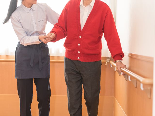 笑顔で手をつなぐ二人 日常風景 スタッフが明るい笑顔でお年寄りと手をつないで歩いています。温かみのある雰囲気が伝わる場面です。