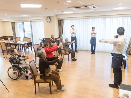 施設の写真 共有スペースに集まり体操をする高齢者と指導をするスタッフ