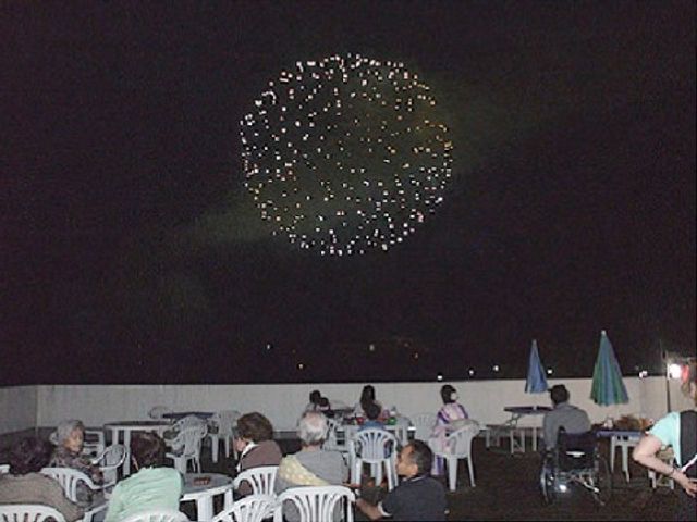 夜空の花火と見物人 花火 夜に開放的なテラスで花火を楽しむ人々の姿が見て取れます。共に特別な瞬間を共有できる場所があります。