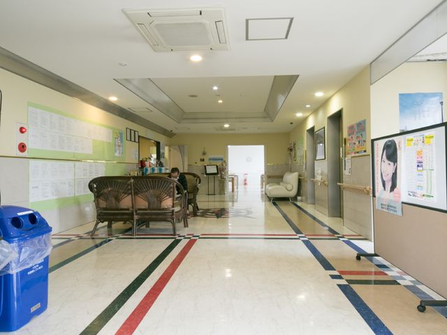 廊下の華やかな情景 広いロビー 清潔感のあふれる廊下には、様々な掲示物があり、利用者とスタッフの穏やかな交流が垣間見えます。ゆったりとした椅子が配置され、くつろぎの空間を演出しています。
