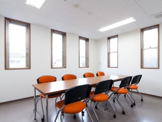 白を基調とした空間には細長い窓がいくつもある。中央に長テーブルがあり、その周りにはオレンジ色の椅子が８脚置かれている。