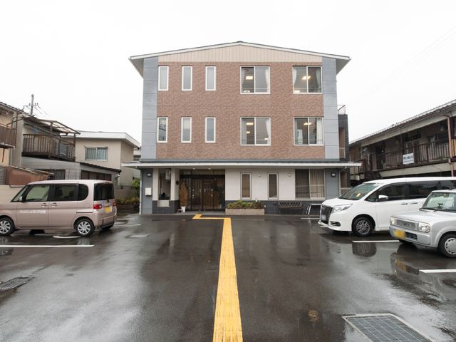 施設前の駐車場 外観1 落ち着いた色合いの建物前には、来訪者用の駐車スペースがあり、アクセスのしやすさが保たれています。雨の日でもスムーズな移動をサポートする設備が充実しており、働く環境に寄り添った配慮が感じられます。
