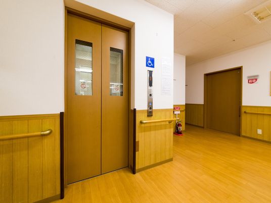 エレベーターの扉は、明るい色のフローリングや建具のイメージと統一された木製である。扉に手を挟まないよう注意喚起するシールが貼られている。