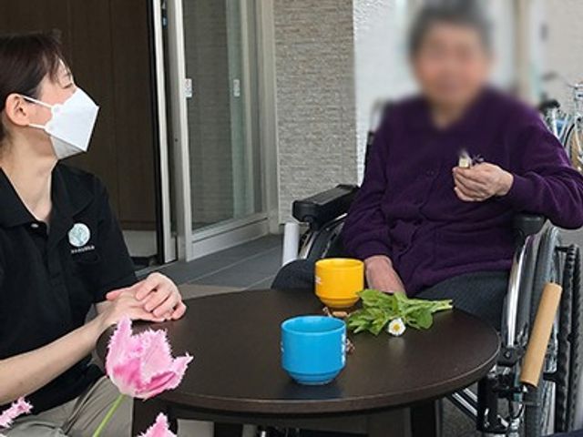 車椅子の居住者とスタッフ テラス 自然光が降り注ぐテラスで、スタッフと居住者が会話を楽しんでいます。リラックスした雰囲気です。