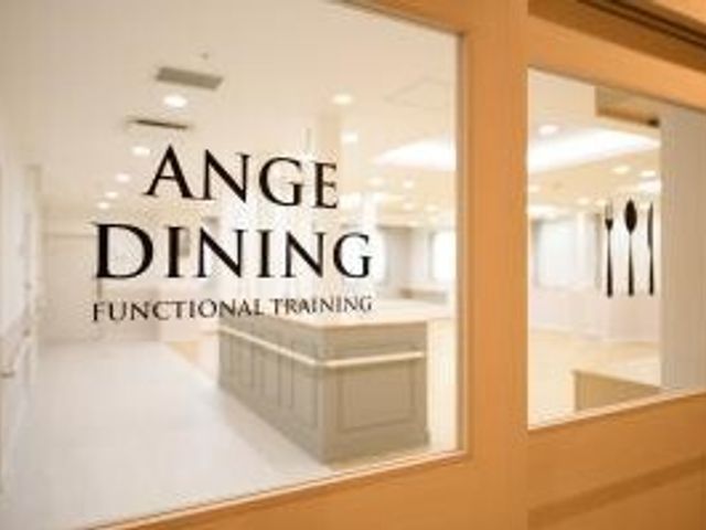 ダイニングとトレーニング室 食堂 アンジュダイニングと表記された食堂の扉越しに見える内装はシンプルで清潔感があります。機能訓練室も併設され、利用者の健康維持に配慮した設計となっています。