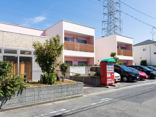 ピンクの建物と青空 外観 明るい青空のもと、落ち着いた色合いの建物が道路に面して立っています。窓が多く、開放感が感じられる設計になっております。