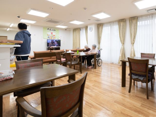 共有スペースの日常風景 リビングダイニング 床の木目が温もりを感じさせる食堂で、利用者の方々が穏やかな時間を過ごしています。スタッフがサポートしながら、日々の生活が営まれています。