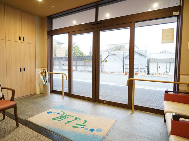 出入口と待合スペース 玄関 ゆとりのあるエントランスは、通路も広くて明るい印象です。待合の椅子が設置されており、訪問者を暖かく迎え入れる空間となっています。