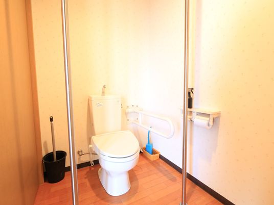 施設の写真 個室トイレの図である。天井まで届くステンレス製のポールが２本あり、その間に便座がある。床に掃除用具が置かれている。