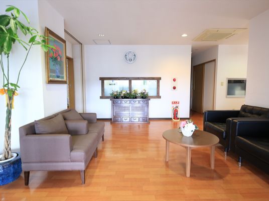 施設の写真 左手にグレーのソファが置かれ、その手前に観葉植物が飾られている。右側の壁には黒いソファがあり、円卓がある。