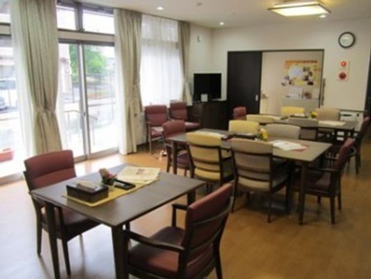 入口付近にテレビがあり、テーブルが３台とそれぞれに椅子が設置されている。レースが束ねられている。窓は大きく採光がいい。