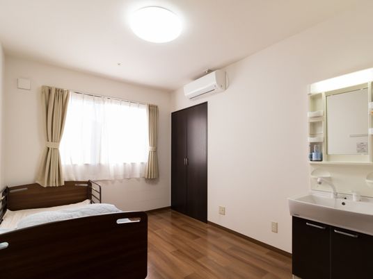 白い壁の中に、焦げ茶色のクローゼットや洗面台、ベッドが設置され、おしゃれな雰囲気である。ベッドの前には広いスペースがある。