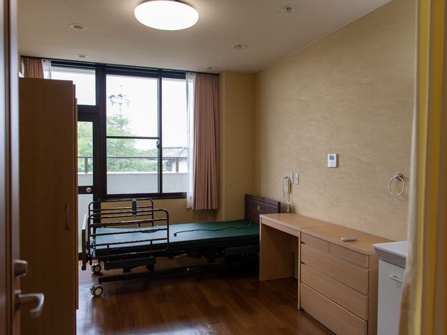 明るい居室の一角 居室 落ち着いた色合いの居室には必要な設備が整っており、窓からは自然光が差し込んでいます。デスクと収納も完備しており、居心地の良い個々のスペースを提供しています。