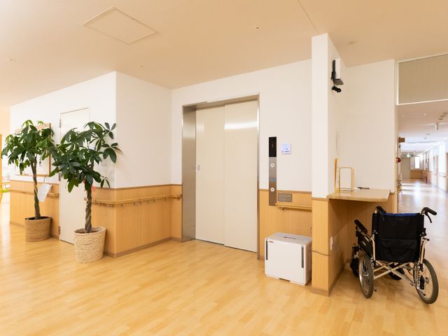 廊下とエレベーター エレベーター 床材が木目調で、清潔感のある廊下が広がっており、車椅子が置かれています。スムーズな移動が考慮された設計になっていることが伺えます。