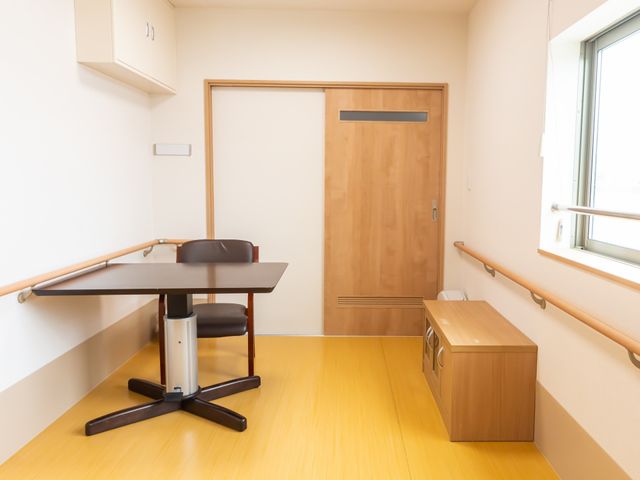 居室のテーブルと扉 休憩室 ゆとりのある単身用居室で、明るい光が差し込み、安心して生活を送れます。壁には手すりが設置され、日々の動作をサポートします。