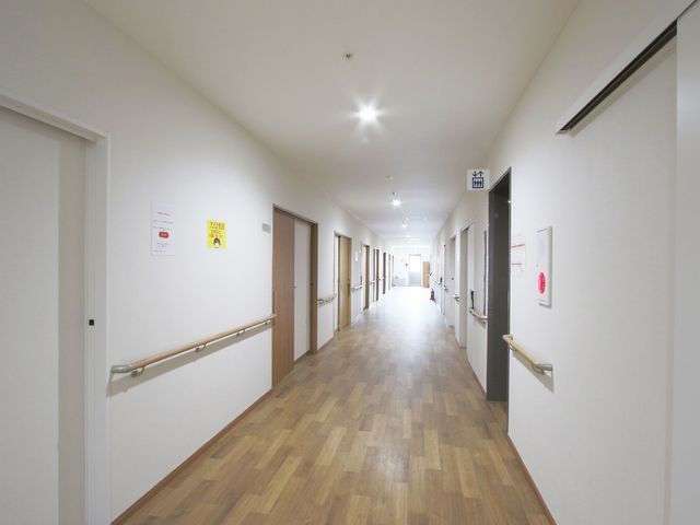 明るく広い施設の廊下 廊下 床に木目調の素材を使用した明るく広々とした廊下です。壁には手すりが備えられ、安全に配慮された設計となっています。