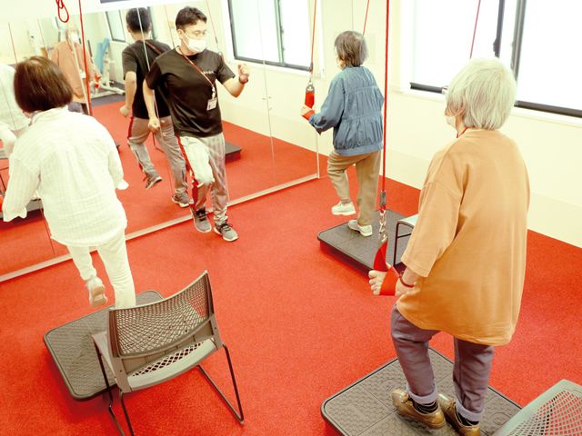 明るい機能訓練室 スタッフの指導のもと、利用者がバランスボードで運動を行っている光景です。日々のリハビリに励む姿が伝わる空間となっています。
